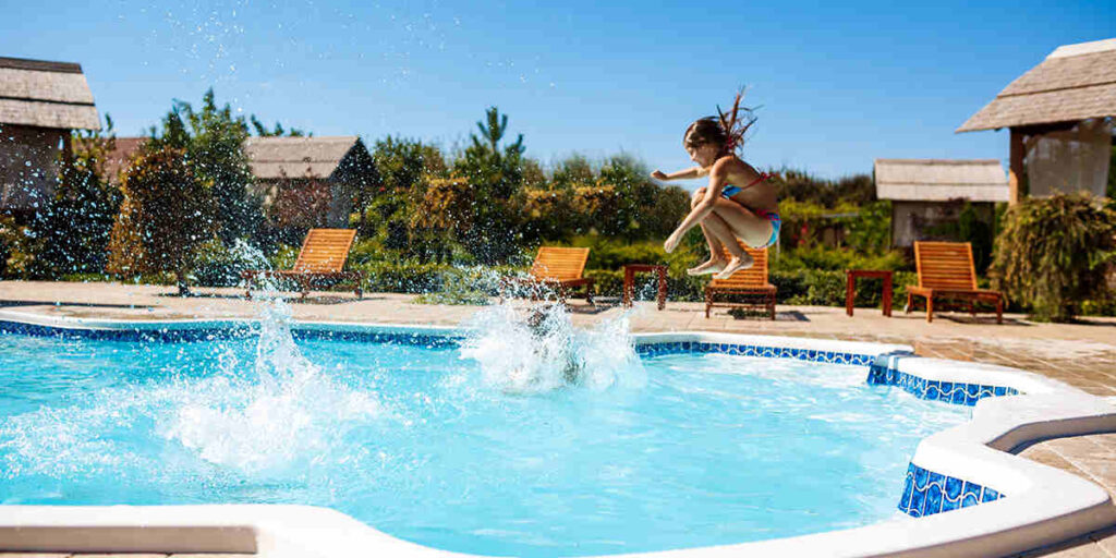 A girl in a swimsuit leaps into a bright blue pool, spraying water as she jumps under a clear sunny sky by poolside loungers and huts.