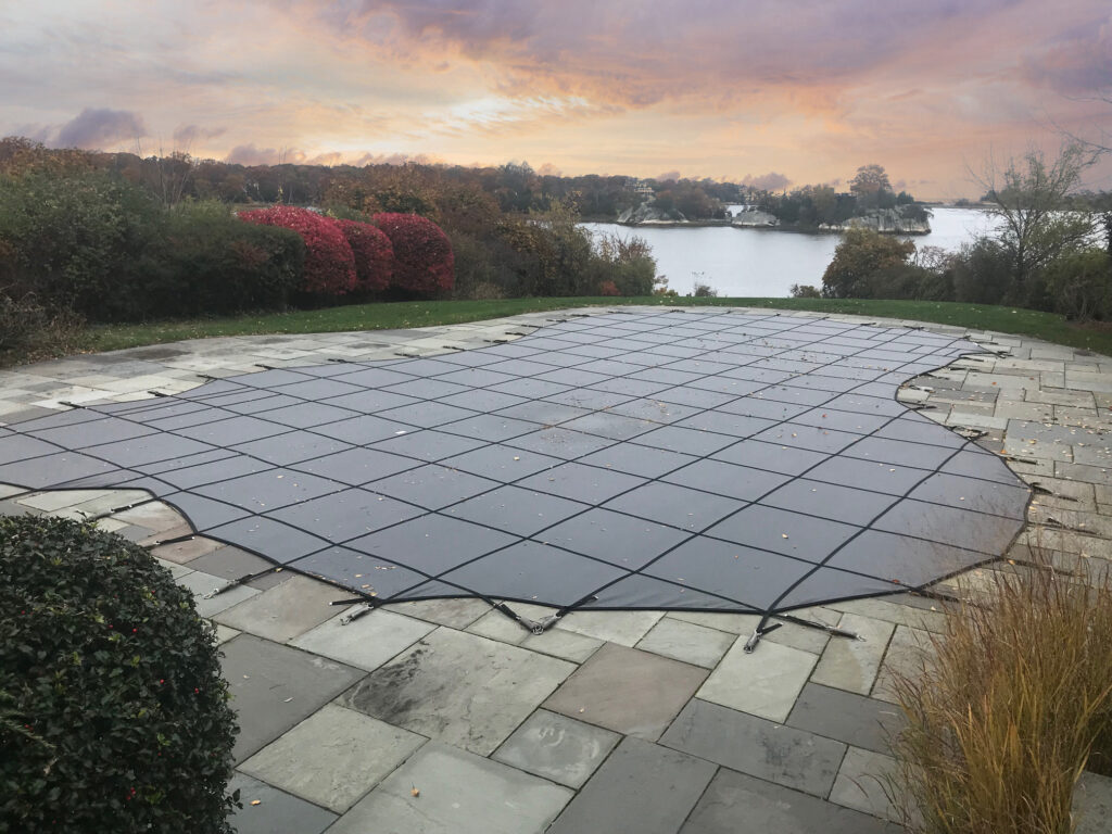 GLI ProMesh Safety Cover | Dickson Family Pools & More Large gray pool GLI ProMesh Safety Cover stretched over a pool, bordered by stone paving and autumn shrubs, with a river and small islands in the distance at sunset.