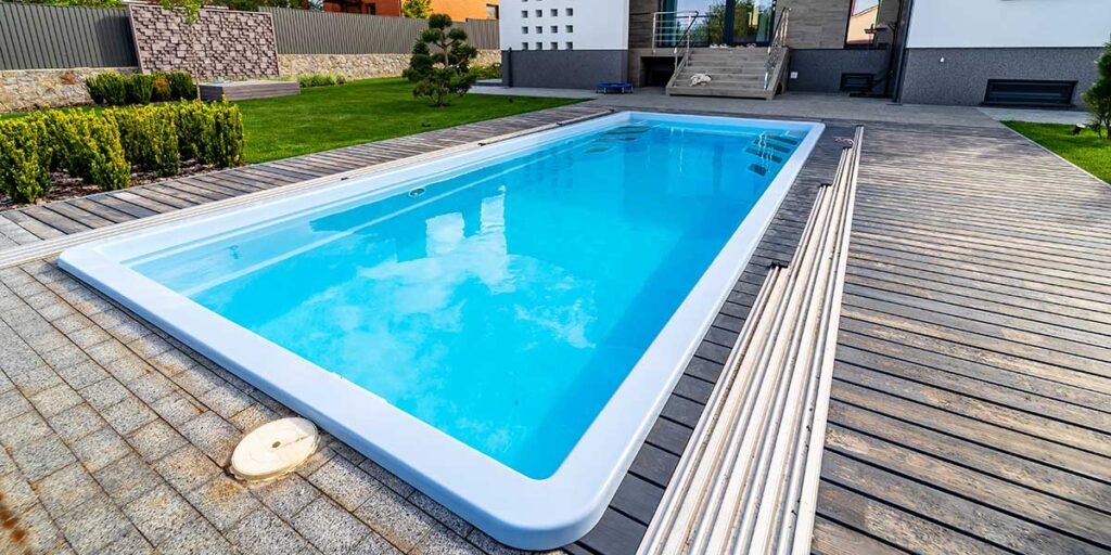 Compact rectangular fiberglass pool installed in a small backyard lawn surrounded by sleek wooden decking and modern stone patio.