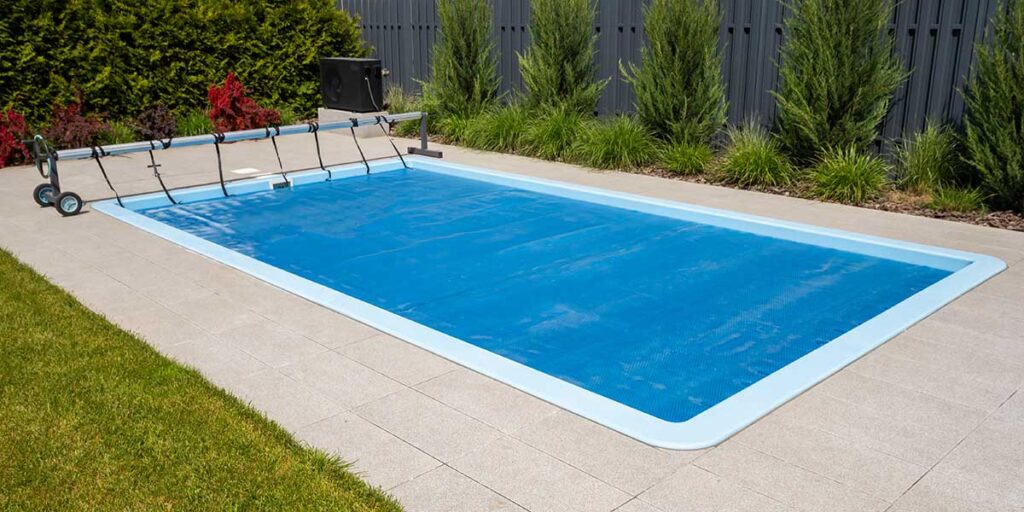 A rectangular backyard vinyl pool covered with a blue protective solar blanket, surrounded by light gray patio pavers.